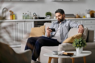 Normal gömlekli ve pantolonlu sakallı bir adam ev ortamında bir kanepede rahatlar, akıllı telefonunu kullanır..