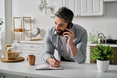 Günlük kıyafetli sakallı bir adam evde defterine not tutarken telefonla konuşuyor..