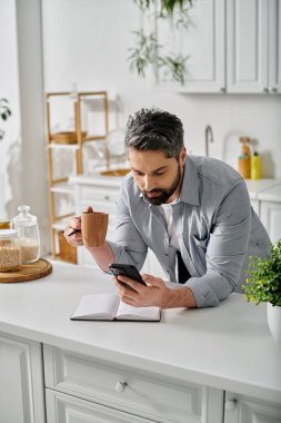 Günlük giysiler içinde sakallı bir adam mutfağında durmuş, bir fincan kahvenin keyfini çıkarıyor.