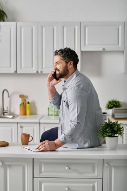 Günlük giysiler içinde sakallı bir adam mutfak tezgahında oturur, telefonuyla konuşur ve not alır..