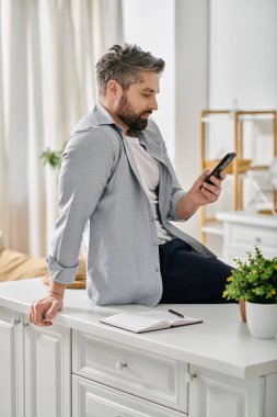 Açık mavi gömlekli sakallı bir adam beyaz mutfak tezgahında otururken telefonunu kontrol ediyor..