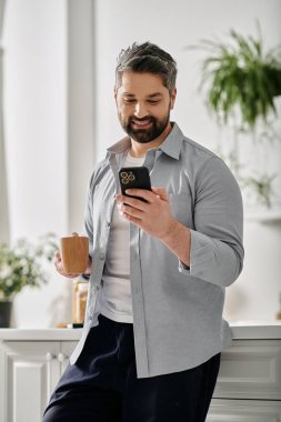 Günlük giysiler içinde sakallı bir adam elinde bir kupa kahve tutarken telefonuna bakıp gülümsüyor..