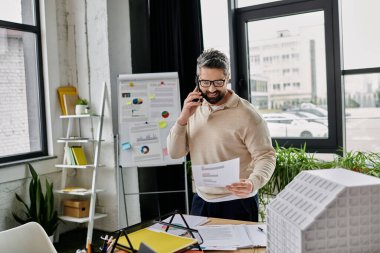 A handsome businessman with a beard stands in a modern office, reviewing plans while speaking on a phone. clipart