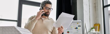 A handsome businessman with a beard works in a modern office, reviewing documents while on a phone call. clipart