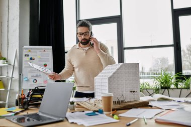 Sakallı yakışıklı bir işadamı, modern bir ofiste telefonla bina planları üzerinde çalışıyor..