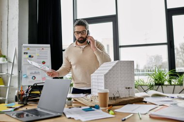 A handsome businessman with a beard works on building plans while on the phone in a modern office. clipart
