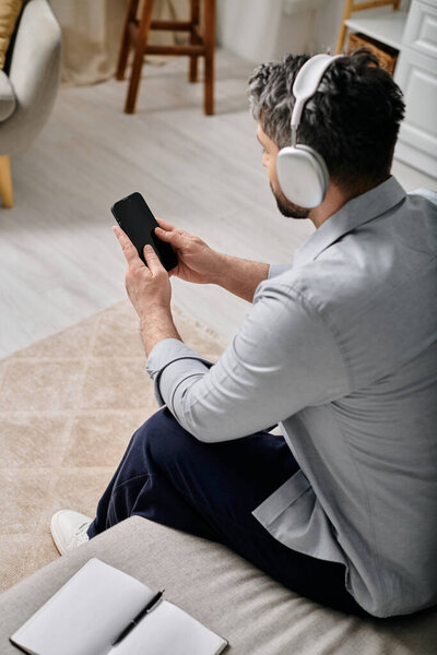 A bearded man wearing headphones sits on a couch, using his smartphone