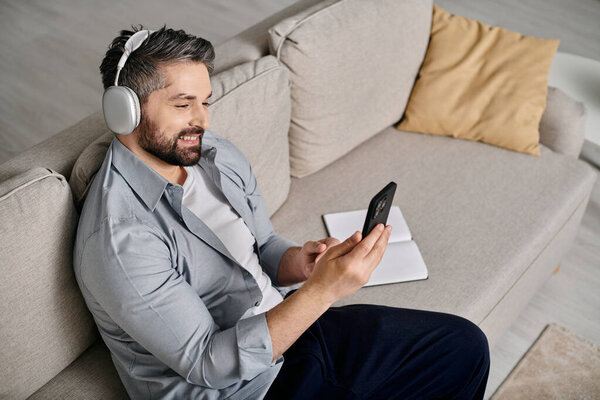 A bearded man in casual attire works remotely from home on his couch, wearing headphones.