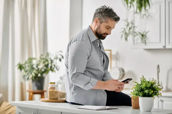 stock image A bearded man in a casual shirt using his phone while sitting at his kitchen counter.