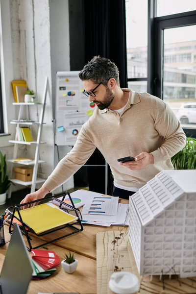A handsome businessman with a beard is working in a modern office. He is organizing papers on his desk while holding a smartphone.