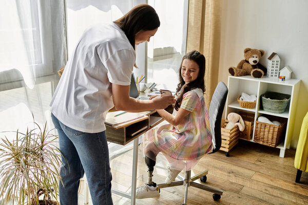 A mother and daughter spend quality time together in their home. The young girl, who has a prosthetic leg, sits at a desk while her mother stands beside her.