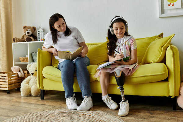 A brunette mother and her daughter with a prosthetic leg are spending quality time together on a yellow sofa in their home.