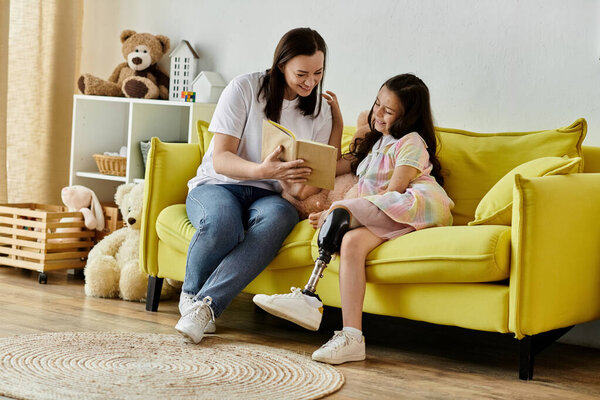 A brunette mother and her daughter, who has a prosthetic leg, are sitting on a yellow couch reading a book together.