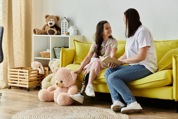 A brunette mother and her daughter, who has a prosthetic leg, are sitting on a yellow couch at home. They are spending quality time together reading a book.