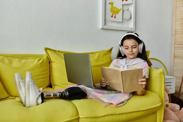 stock image A young girl with a prosthetic leg relaxes on a yellow sofa, lost in a book while wearing headphones.