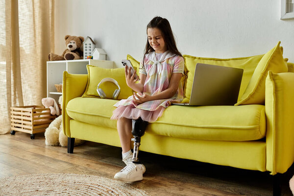 A young girl with a prosthetic leg smiles while using her phone, sitting on a yellow sofa.