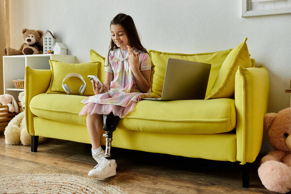 A young girl with a prosthetic leg sits on a yellow sofa, smiling and holding a phone.