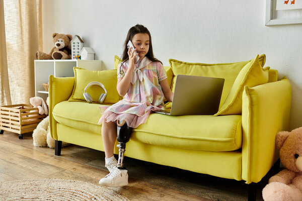 A young girl with a prosthetic leg sits on a yellow couch, using a laptop and talking on a phone.