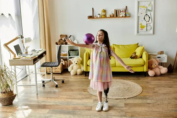 stock image A young girl with a prosthetic leg throws a ball while smiling in her home, showcasing her playful spirit and determination.