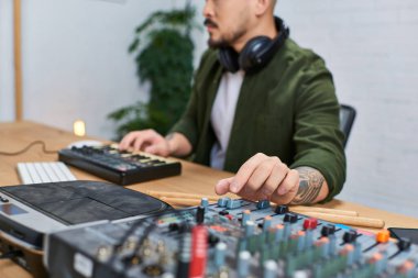 Asyalı bir adam müzik ekipmanlarıyla çevrili atölyesinde oturuyor ve sanatına odaklanıyor..