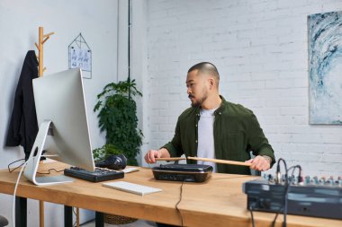 Yakışıklı Asyalı bir adam, günlük kıyafetler giymiş, stüdyosunda oturuyor ve elektronik bir bateri platformunda davul çalıyor..
