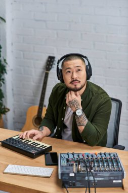 Kulaklık takan Asyalı bir adam stüdyosunda oturuyor ve klavye çalarken düşünceli görünüyor..