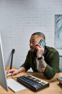 Yakışıklı bir Asyalı, muhtemelen çok enstrümanlı bir adam, stüdyosunda müzik üzerine çalışıyor, klavye ve bilgisayar kullanıyor. Telefonla konuşuyor ve işine odaklanmış gibi görünüyor..