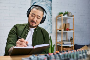 Sıradan giyinmiş genç bir Asyalı bir adam bir müzik stüdyosunda kulaklık takarak bir deftere yazıyor..