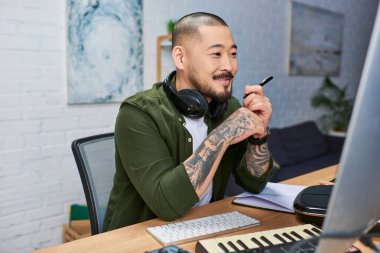 Kulaklık takan yakışıklı Asyalı bir adam masasına oturmuş elinde bir kalem tutarak bilgisayar ekranına dikkatlice bakıyor. Stüdyosunda müzik bestelemeye odaklanmış gibi görünüyor..