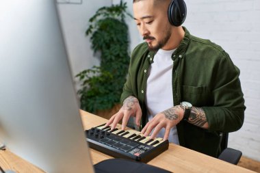 A young Asian man wearing headphones, a green shirt and a white undershirt plays on a midi keyboard in his studio. clipart