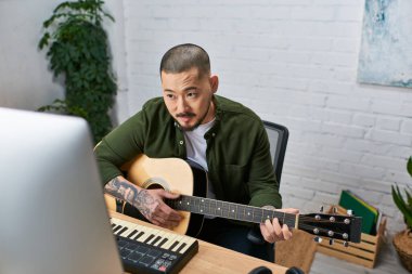 Bir adam stüdyosunda bir bilgisayar ekranına bakarken gitar çalar..