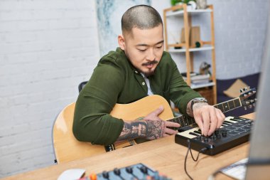 Dövmeli yakışıklı bir Asyalı akustik gitar ve müzik stüdyosunda MIDI klavye çalıyor..