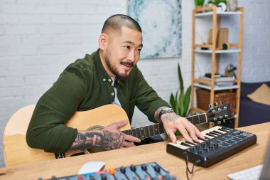 Yakışıklı Asyalı bir adam stüdyosunda müzik yaparken gitar ve klavye çalıyor..