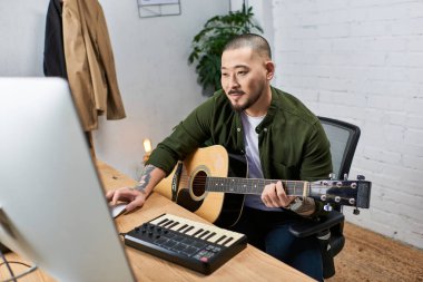 Genç bir Asyalı akustik gitar çalıyor ve stüdyosunda klavye kullanıyor..