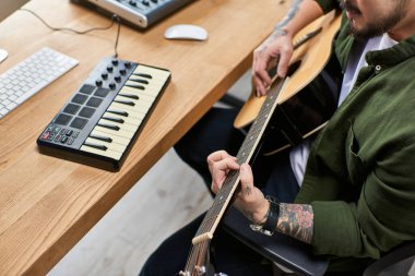 A handsome Asian man plays an acoustic guitar in his studio, surrounded by musical equipment. clipart