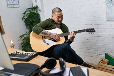 A handsome Asian man plays acoustic guitar in his studio. clipart