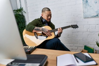 Genç bir Asyalı, müzik aletleri ve ekipmanlarıyla çevrili olan stüdyosunda akustik gitar çalıyor..