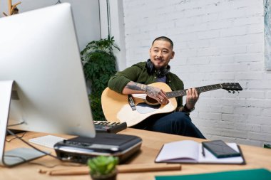 A handsome Asian man with tattoos sits in a studio, playing an acoustic guitar. clipart