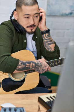 Günlük kıyafetlerle dövmeli Asyalı bir adam stüdyosunda kulaklık takarken akustik gitar çalıyor..