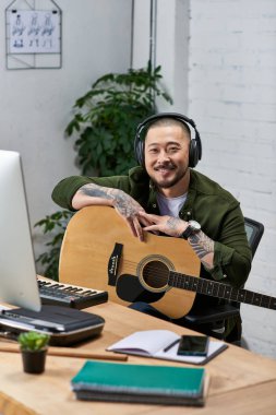 Yakışıklı bir Asyalı stüdyosunda gitar ve kulaklıkla otururken gülümsüyor..