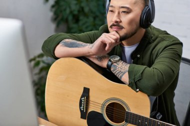 Yakışıklı Asyalı bir adam, çok enstrümanlı bir adam, günlük giysiler içinde, bir stüdyoda gitarına yaslanıyor..