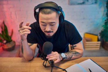 An Asian man sits in his home studio, wearing headphones and speaking into a microphone. clipart