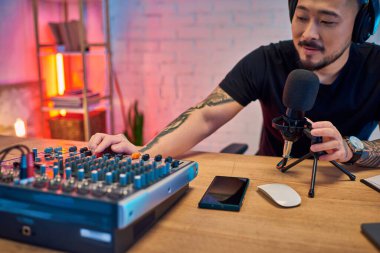 Asyalı bir adam stüdyosunda mikrofon ve ses karıştırıcı kullanarak bir podcast kaydediyor..