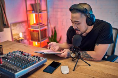 Adamın biri, önünde mikrofon, ses karıştırıcı ve defter bulunan bir podcast 'i kendi stüdyosunda kaydediyor..