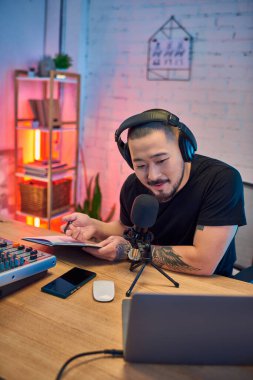 Genç Asyalı bir adam stüdyosunda bir podcast kaydediyor, kulaklık takıyor ve mikrofona konuşuyor..