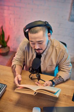Genç bir Asyalı podcast stüdyosunda oturuyor, kulaklıkları açık, elinde açık bir kitapla bir bölüm kaydediyor..
