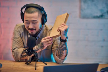 Asyalı bir adam stüdyosunda oturmuş, kulaklık takıyor ve elinde kitap tutarken podcast yapıyor..
