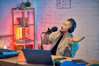 Asyalı bir adam stüdyo ortamında bir podcast kaydediyor, mikrofon tutuyor ve kulaklık takıp gülümsüyor..