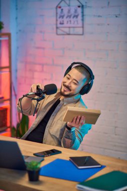 Yakışıklı bir Asyalı elinde mikrofonuyla podcast 'ini kaydederken gülümsüyor. Renkli stüdyo ışıklarıyla çevrili..