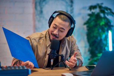 Kulaklıklı yakışıklı bir Asyalı stüdyoda bir podcast kaydediyor, elinde mavi bir kağıt tutarak mikrofona konuşuyor..